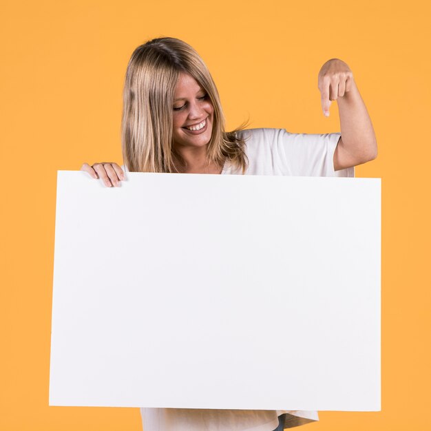 Mujer joven sonriente que señala el dedo índice en el cartel en blanco blanco
