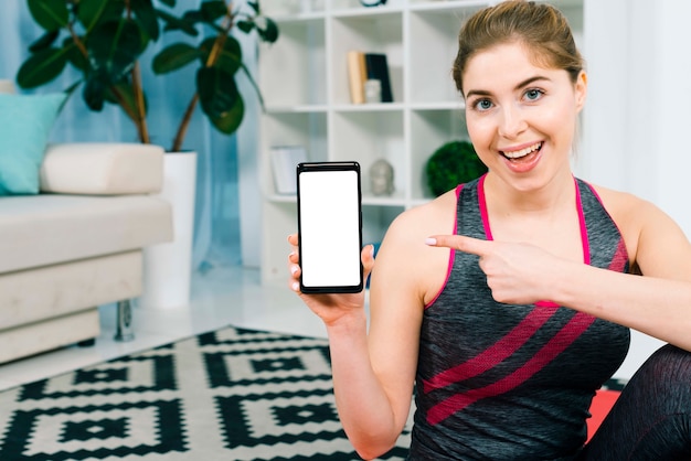 Foto gratuita mujer joven sonriente que señala su dedo hacia su teléfono móvil que se sienta en la sala de estar