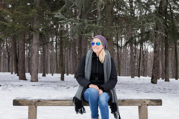 Mujer joven sonriente que se sienta en banco de madera en invierno en el bosque nevoso