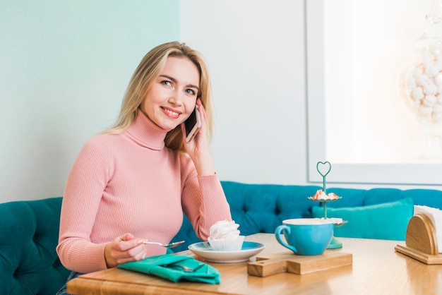 Foto gratuita mujer joven sonriente que se sienta en el café que habla en el teléfono móvil que goza de la magdalena y del café