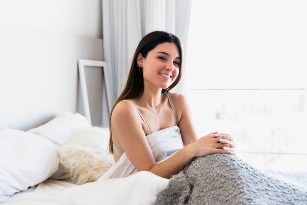 Mujer joven sonriente que se sienta en cama en el dormitorio