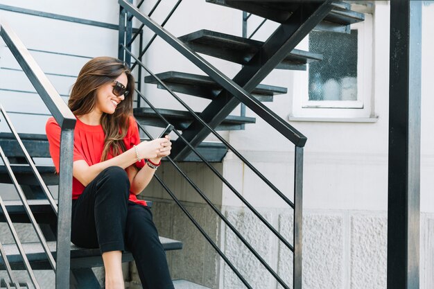 Mujer joven sonriente que se sienta en la escalera usando el teléfono móvil