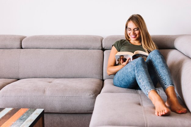Mujer joven sonriente que se sienta en el libro de lectura del sofá