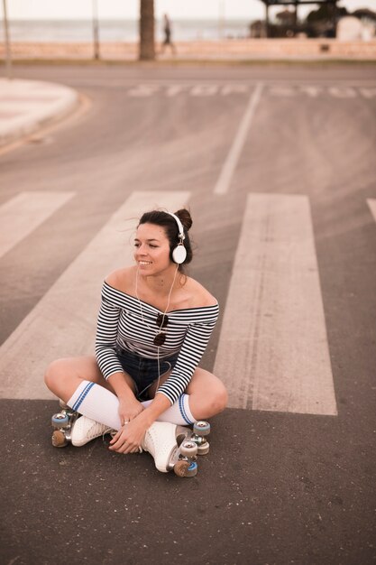 Mujer joven sonriente que se sienta en música que escucha del patín de ruedas del camino que lleva en el auricular