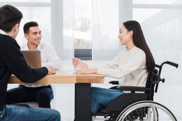 Mujer joven sonriente que se sienta en la silla de ruedas que tiene reunión de negocios corporativa con sus dos colega