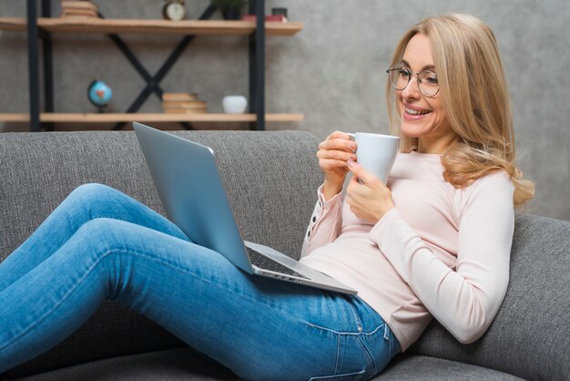 Mujer joven sonriente que se sienta en el sofá que sostiene la taza de café que mira el ordenador portátil