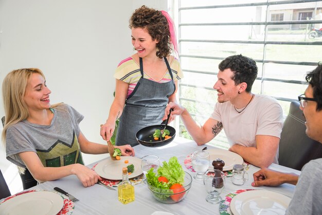 Mujer joven sonriente que sirve verduras cocinadas a sus amigos en la mesa de comedor