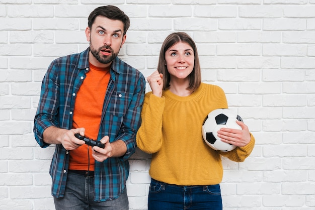 Foto gratuita mujer joven sonriente que sostiene el balón de fútbol en la mano que anima a su novio que juega al videojuego