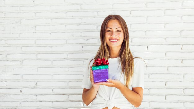 Mujer joven sonriente que sostiene la caja de regalo delante de la pared de ladrillo