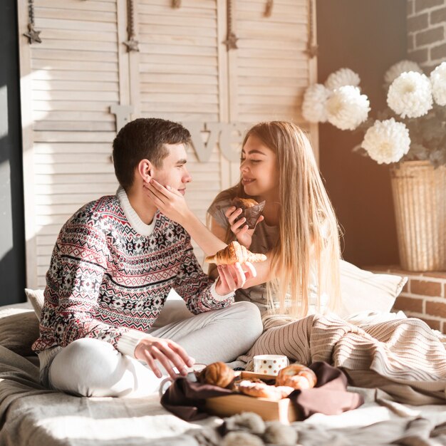 Mujer joven sonriente que sostiene la magdalena que ama a su novio que sostiene el cruasán