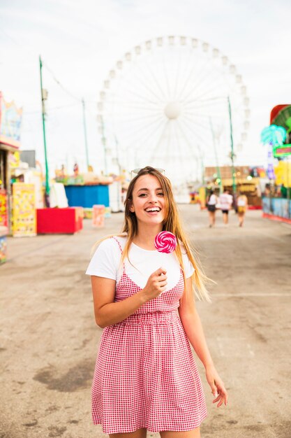 Mujer joven sonriente que sostiene la piruleta en la mano que se coloca en el parque de atracciones