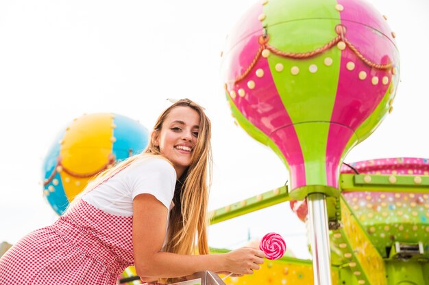 Mujer joven sonriente que sostiene la piruleta en el parque de atracciones