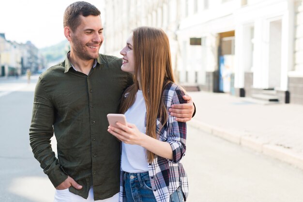 Mujer joven sonriente que sostiene smartphone y que mira a su novio en la calle