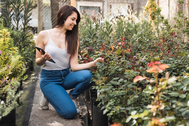 Mujer joven sonriente que sostiene la tableta digital disponible que cultiva un huerto en el invernadero