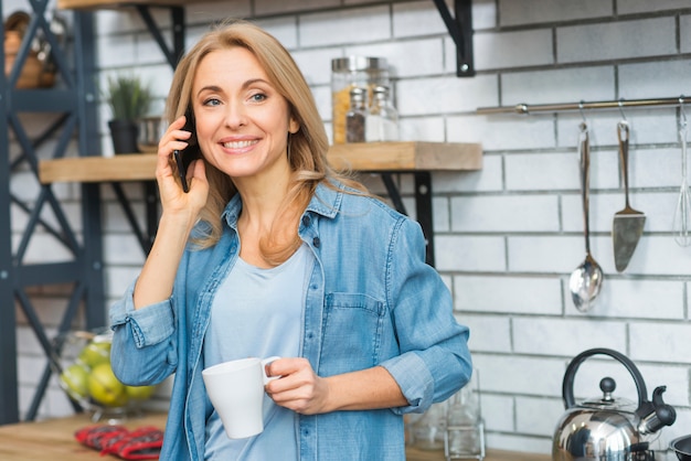 Foto gratuita mujer joven sonriente que sostiene la taza blanca disponible que habla en el teléfono móvil