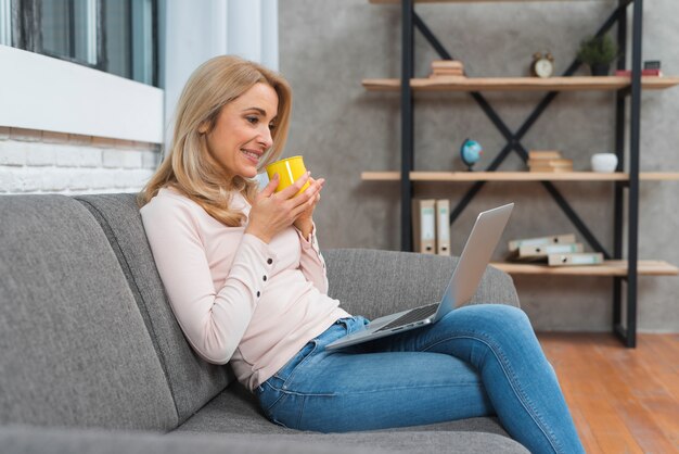 Mujer joven sonriente que sostiene la taza de café que mira el ordenador portátil