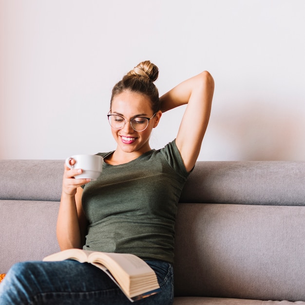 Mujer joven sonriente que sostiene la taza de café que se sienta en el libro de lectura del sofá