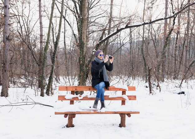 Mujer joven sonriente que toma las fotografías en el invierno que se sienta en banco en nieve