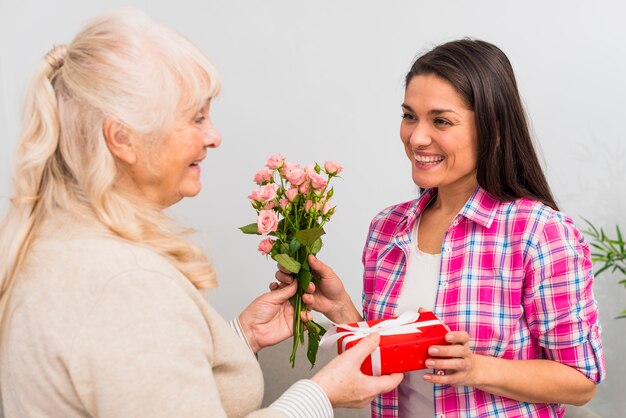 Mujer joven sonriente que toma el regalo y el ramo color de rosa de su mujer mayor