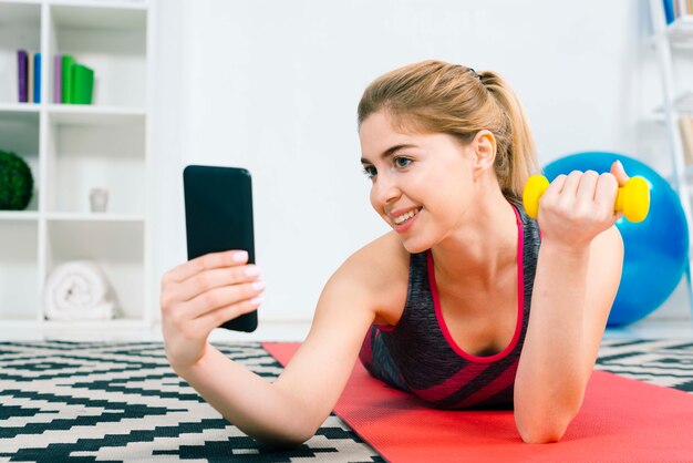 Mujer joven sonriente que toma el selfie en el teléfono móvil mientras que hace ejercicio con pesa de gimnasia amarilla
