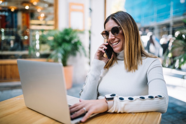 Mujer joven sonriente que usa el ordenador portátil y el smartphone en la tabla en café de la calle