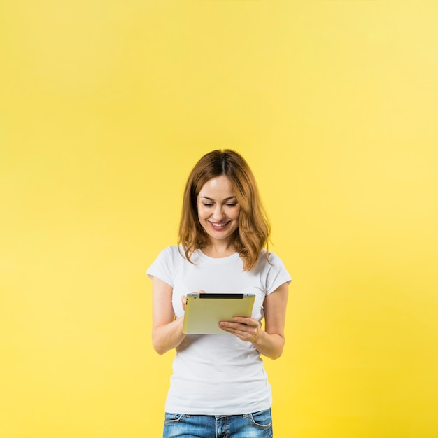 Mujer joven sonriente que usa la tableta digital contra el contexto amarillo