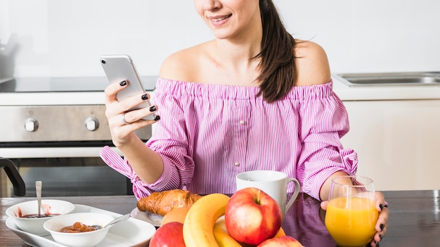 Mujer joven sonriente que usa el teléfono móvil que sostiene el vidrio de jugo