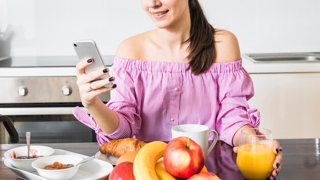 Mujer joven sonriente que usa el teléfono móvil que sostiene el vidrio de jugo