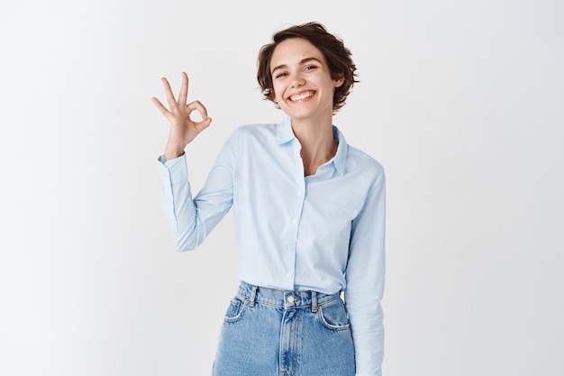 La mujer joven sonriente se siente segura, mostrando un buen gesto de aprobación, elogia el buen producto, de pie en la pared blanca