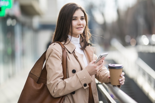 Mujer joven sonriente con la taza de café en el teléfono en la ciudad