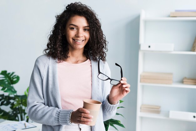 Mujer joven sonriente con la taza de papel y los vidrios en lugar de trabajo