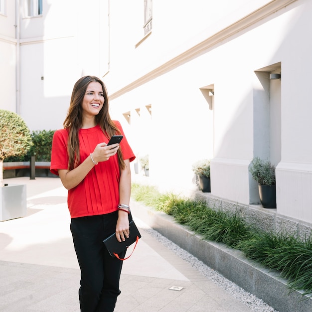 Mujer joven sonriente con el teléfono móvil que se coloca cerca del edificio residencial