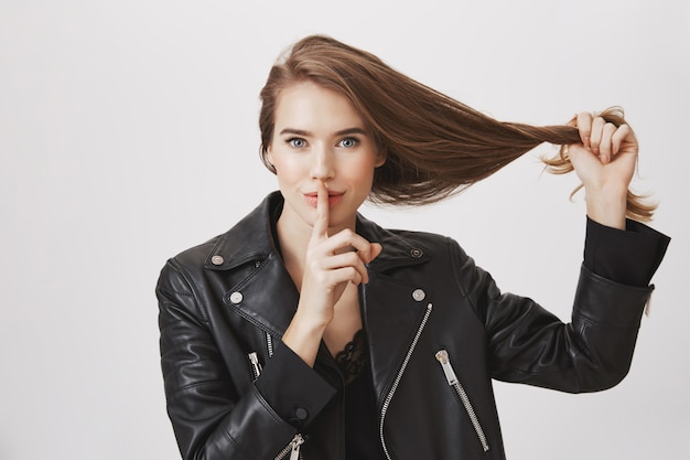 La mujer joven sonriente tira del pelo, hace la señal del silencio, diciendo secreto. Concepto de productos para el cuidado del cabello