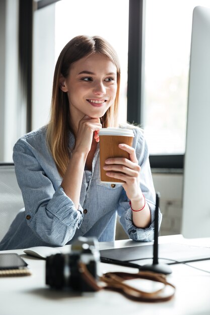 La mujer joven sonriente trabaja en oficina usando la computadora que bebe el café.