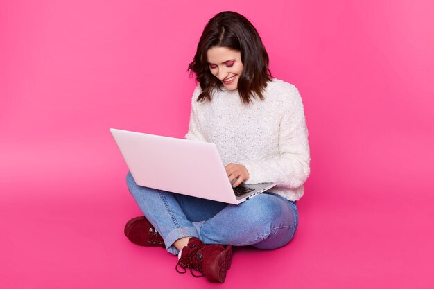 La mujer joven sonriente utiliza la computadora portátil para ganar dinero en Internet. Encantadora morena trabaja en línea, se sienta con las piernas cruzadas en el piso. Lady lleva un suéter blanco informal, jeans y cómodas botas granate.