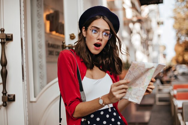 Mujer joven sorprendida con boina elegante, traje blanco y rojo, gafas de pie con mapa en la terraza de la cafetería de la ciudad y mirando asombrado