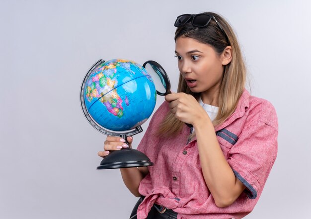 Una mujer joven sorprendida con camisa roja en gafas de sol sosteniendo un globo terráqueo mientras lo mira con lupa