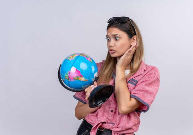 Una mujer joven sorprendida con camisa roja con gafas de sol sosteniendo un globo terráqueo mientras lo mira en una pared blanca