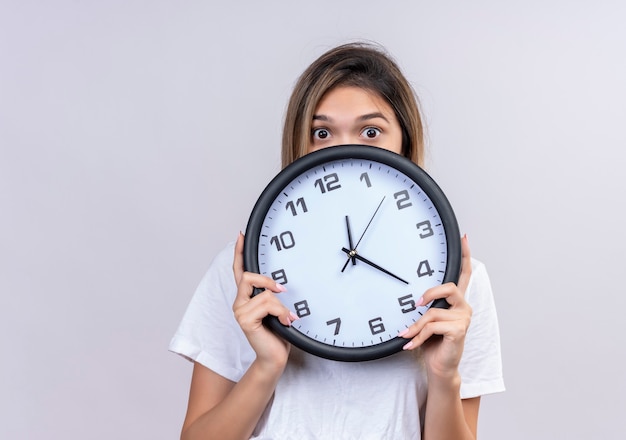 Una mujer joven sorprendida en camiseta blanca sosteniendo el reloj de pared mientras mira