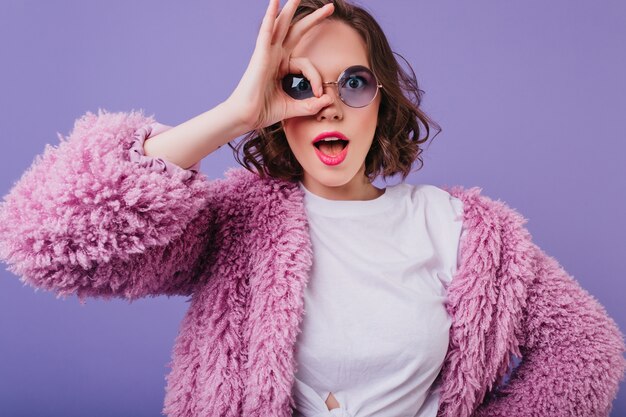 Mujer joven sorprendida en chaqueta mullida haciendo muecas en la pared púrpura. espectacular chica blanca con gafas de sol bromeando.