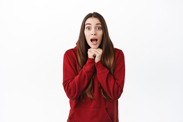 Mujer joven sorprendida escucha la historia con emoción, jadeando y luciendo fascinada, viendo algo sorprendente e interesante, de pie con una sudadera con capucha roja sobre fondo blanco.