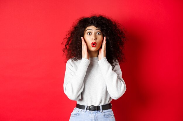 Mujer joven sorprendida escucha noticias increíbles, mirando promo y diciendo wow, mira fijamente con incredulidad, de pie sobre fondo rojo