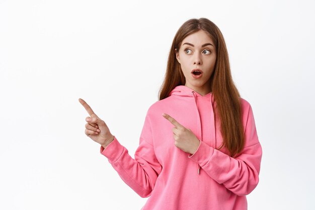 Mujer joven sorprendida jadeando apuntando a la izquierda en copyspace a un lado mostrando un anuncio de pie sobre fondo blanco.