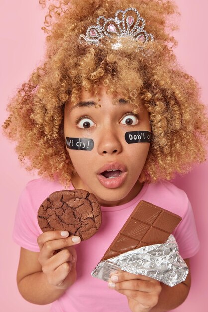 Mujer joven sorprendida mira fijamente a la cámara con ojos saltones sostiene una galleta apetitosa y una barra de chocolate temerosa de obtener un exceso de calorías vestida con una camiseta casual aislada sobre fondo rosa