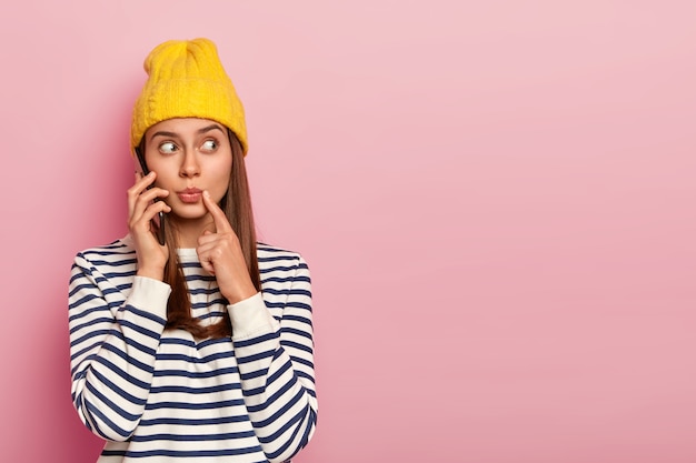 Mujer joven sorprendida mira sorprendentemente a un lado, mantiene el teléfono móvil cerca del oído, tiene una conversación telefónica, mira hacia otro lado, viste un traje de moda