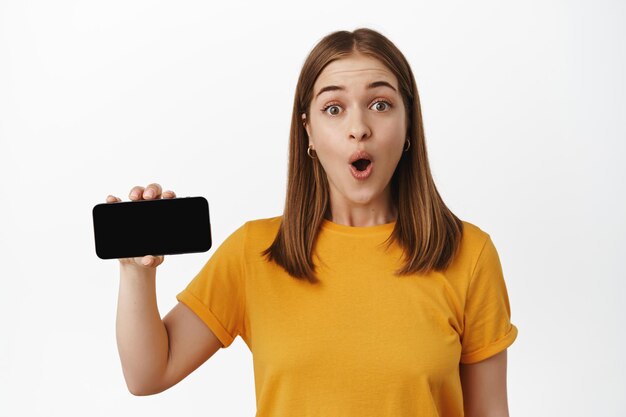 Mujer joven sorprendida muestra la pantalla horizontal del teléfono inteligente, la interfaz de la aplicación, la venta o el sitio de la empresa en el teléfono móvil, mira impresionada a la cámara, camiseta amarilla, fondo blanco