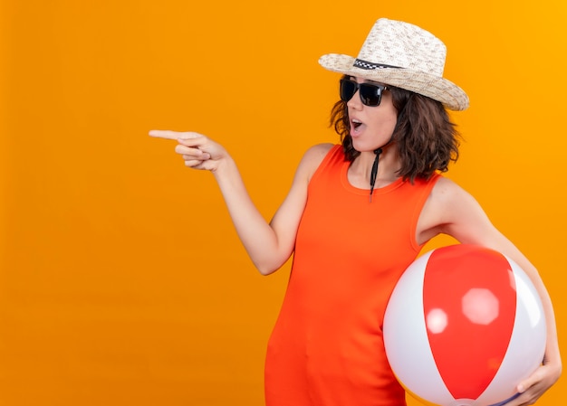 Foto gratuita una mujer joven sorprendida con el pelo corto en una camisa naranja con sombrero para el sol y gafas de sol sosteniendo una bola inflable apuntando al lado derecho con el dedo índice