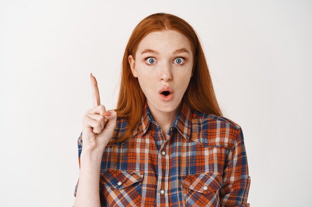 Mujer joven sorprendida con el pelo largo rojo y piel pálida, con aspecto emocionado y apuntando con el dedo hacia arriba, mostrando una oferta de promoción impresionante, de pie sobre una pared blanca