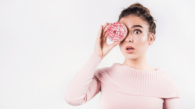 Una mujer joven sorprendida pone mala cara a sus labios cubriendo los ojos con una dona sobre fondo blanco