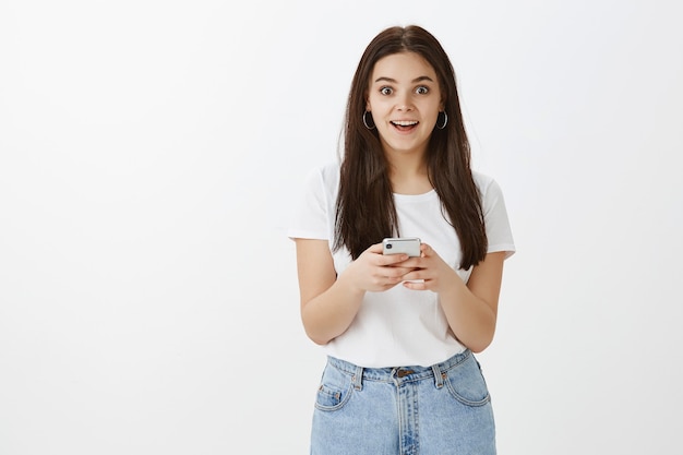 Foto gratuita mujer joven sorprendida posando con su teléfono contra la pared blanca